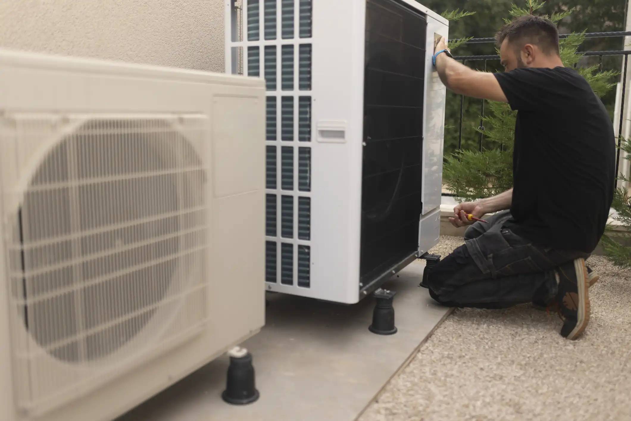 Technician kneels on gravel beside two outdoor HVAC units, using tool to perform maintenance or installation.