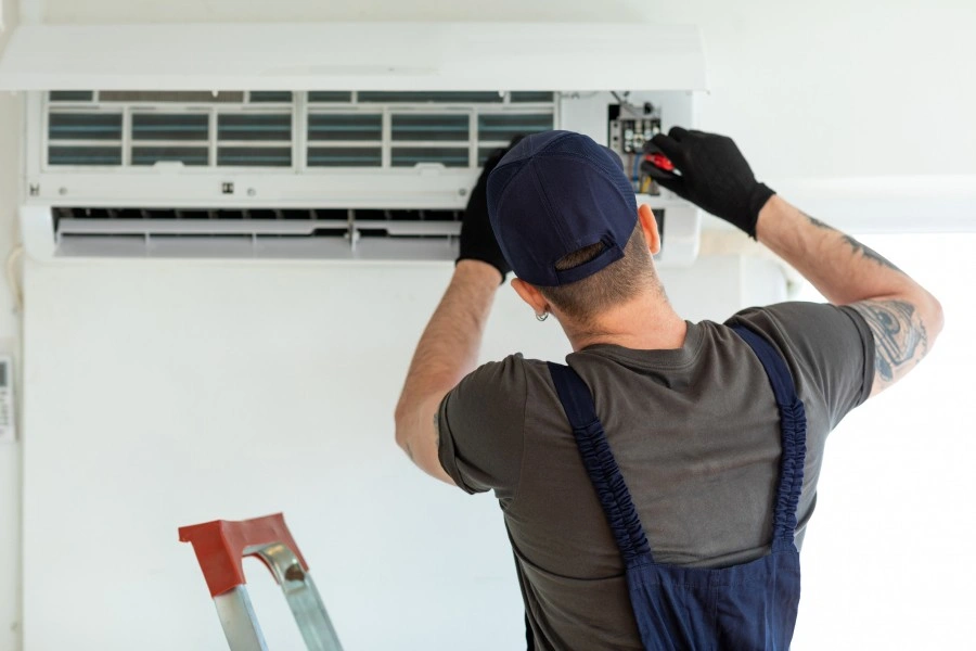 A technician wearing gloves and a cap is working on an air conditioning unit mounted on the wall, with a ladder nearby.