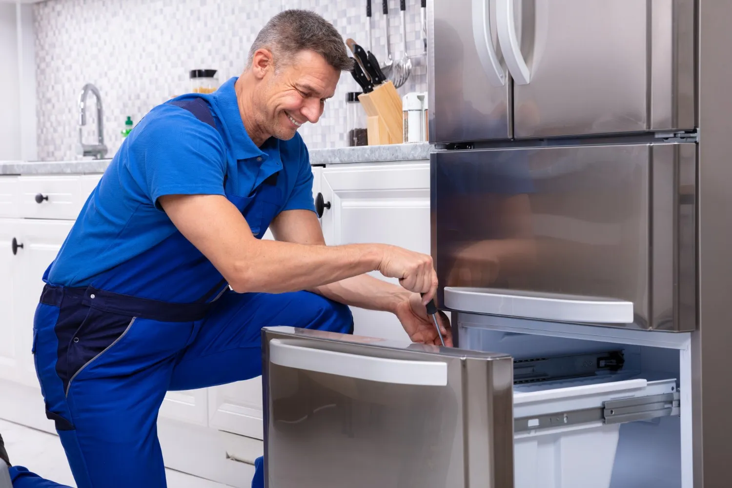 Technician in blue uniform kneels to repair stainless steel refrigerator’s freezer compartment in modern kitchen.