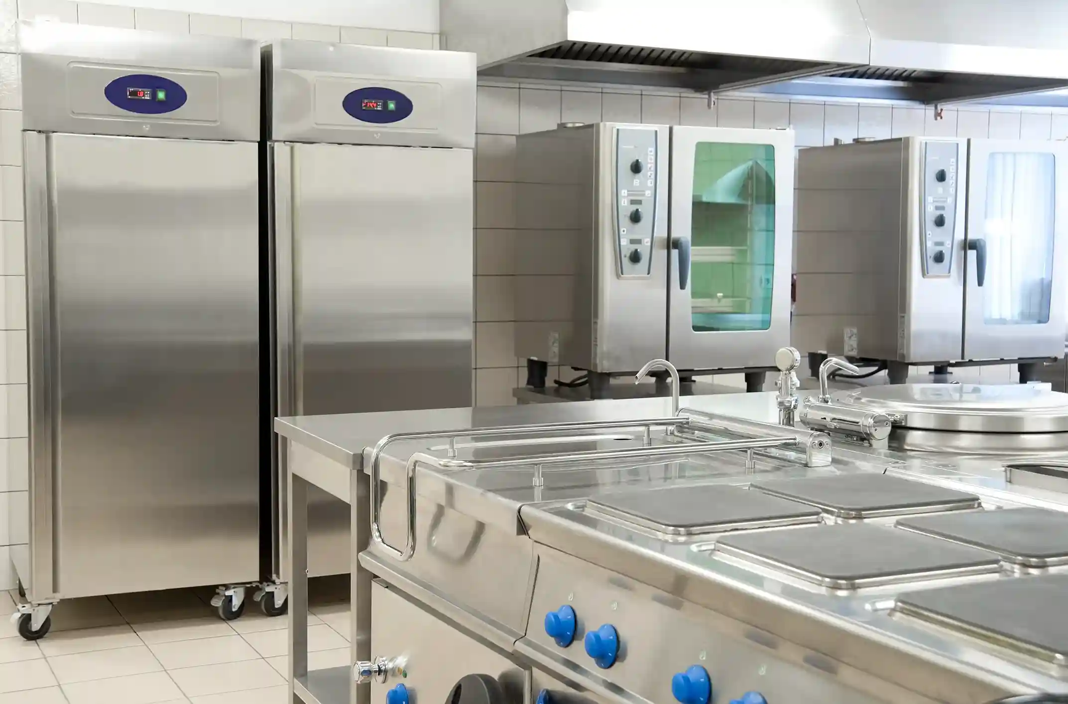 A commercial kitchen with stainless steel appliances, including two large refrigerators on wheels, a stove top, and convection ovens.