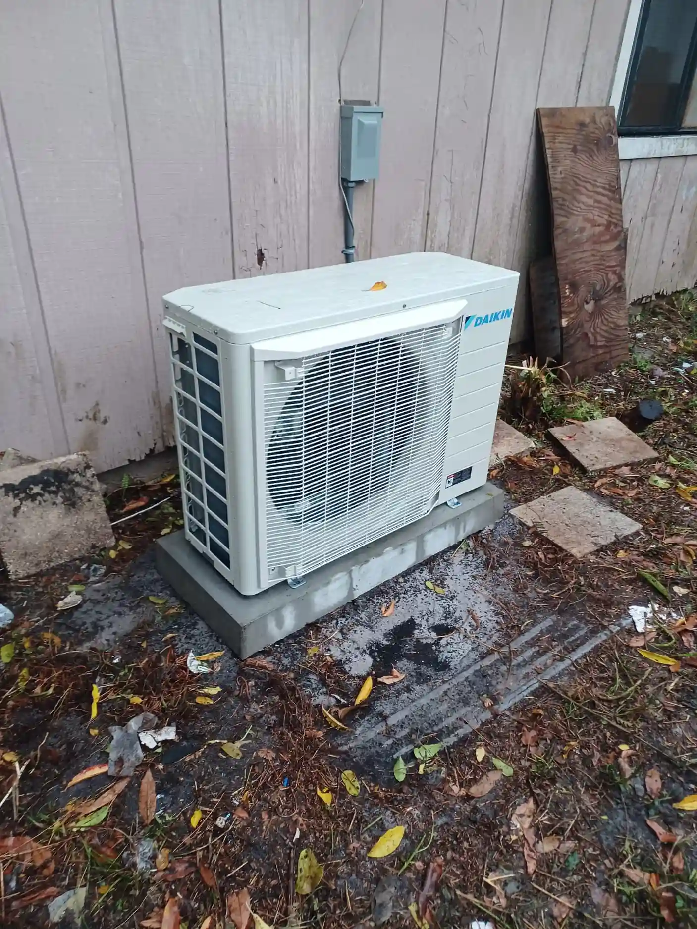 Ductless mini-split outdoor unit installed near a wall, showcasing energy-efficient HVAC technology by Daikin, surrounded by leaves and debris.