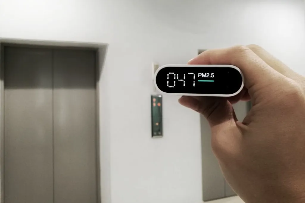 Hand holding air quality monitor displaying PM2.5 level of 47 in front of elevator doors, emphasizing the importance of indoor air quality related to duct efficiency and HVAC systems.