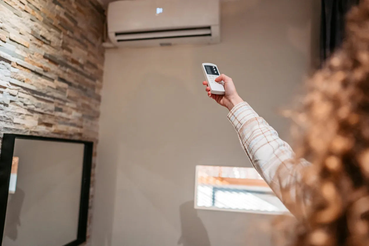 Person using remote control to operate a wall-mounted air conditioning unit in a modern indoor setting, emphasizing HVAC efficiency and air quality improvement.
