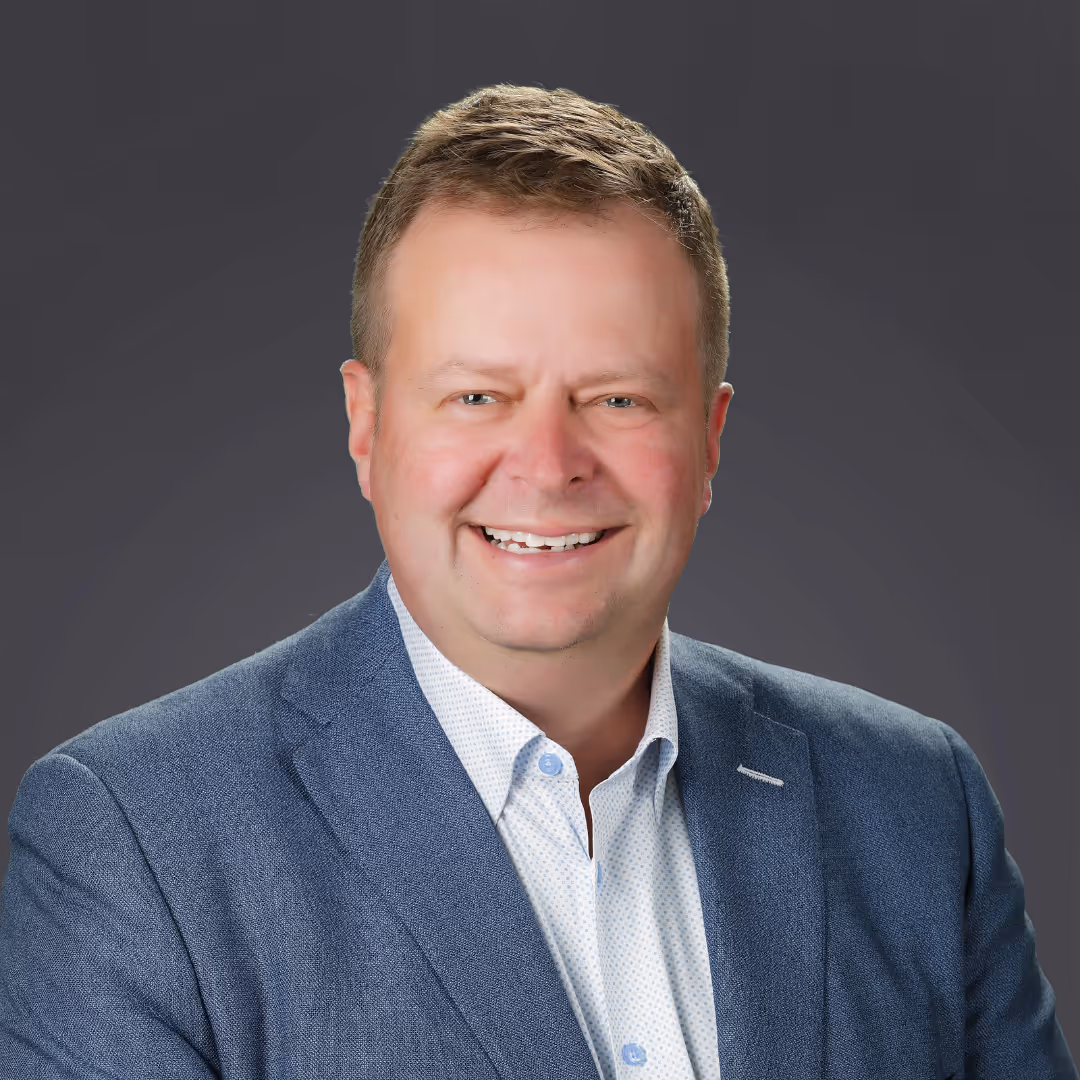 Smiling middle-aged man wearing a blue suit jacket and white dress shirt against a dark gray background.
