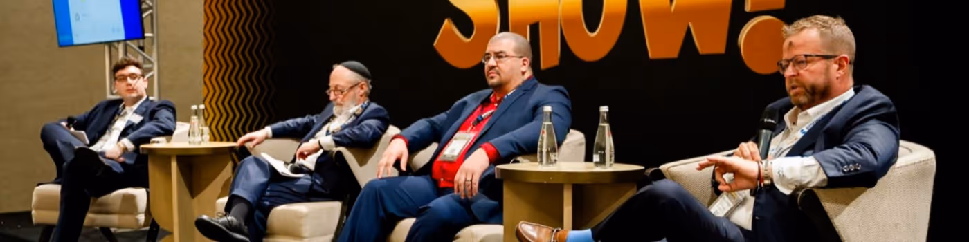 Four men in suits seated on stage chairs during a panel discussion, one speaking into a microphone and checking his watch.