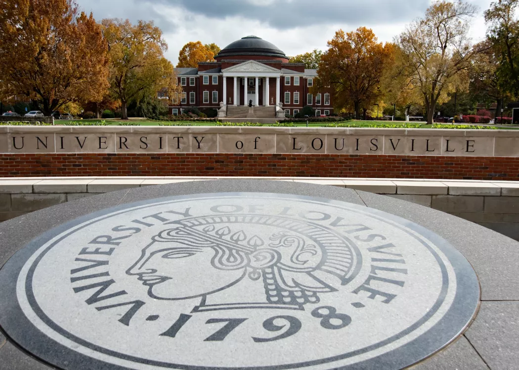 University of Louisville emblem on ground with historic red brick building and autumn trees in background.