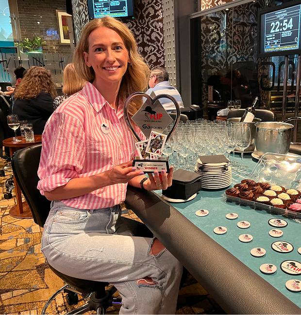Woman holding a poker tournament winner trophy at a casino-style event, seated at a poker table with chips and refreshments.