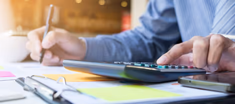 Close-up of a person using a calculator and writing with a pen on documents at a desk.