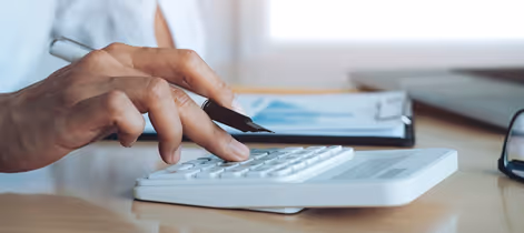 Person using a white calculator with one hand and holding a pen over documents on a desk.