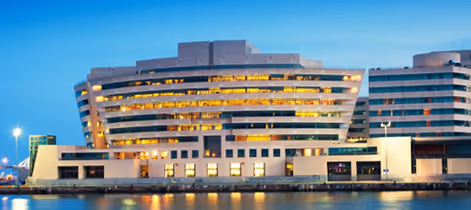 Modern multi-story building illuminated at dusk beside calm water reflecting the lights.