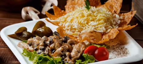 Plate with creamy chicken and mushroom dish on lettuce, cherry tomatoes, pickles, and rice served in a crispy tortilla bowl.