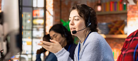 Woman wearing headset speaking and gesturing in a busy call center with colleagues in the background.