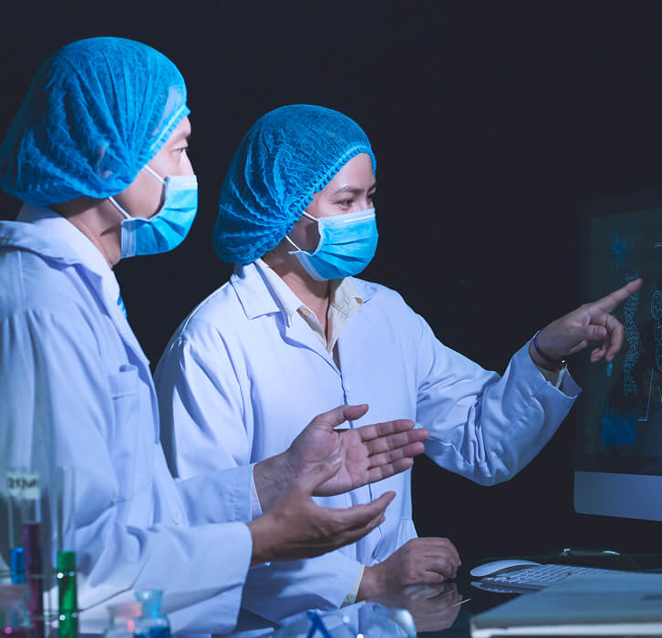 Two scientists in lab coats, masks, and blue hair covers analyzing molecular structure images on a computer screen.