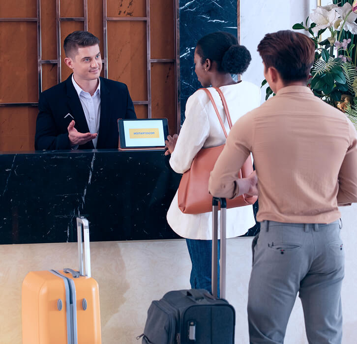 Hotel receptionist talking to two guests with luggage at the front desk.