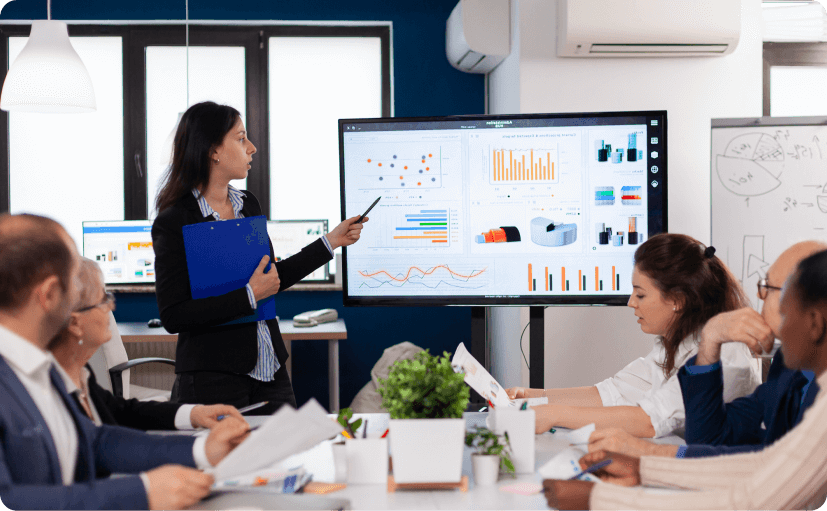 Woman in business attire giving a presentation using a large screen displaying various charts and graphs to a group of attentive colleagues in a modern office.