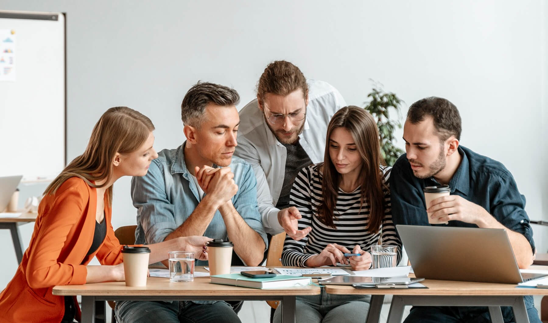Five adults collaborating around a table with documents, laptops, and coffee cups in a bright office.