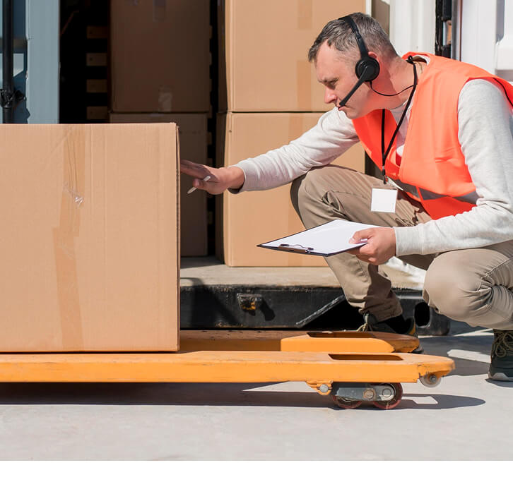 Worker in orange safety vest and headset inspecting a large cardboard box on an orange pallet jack near a delivery truck.