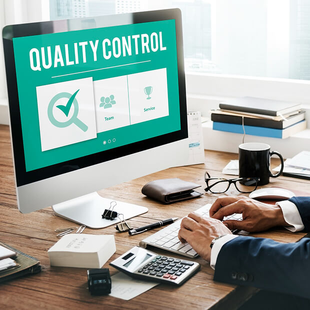 Person in business attire working on a desktop computer displaying a screen with 'Quality Control' and icons labeled Team and Service on a wooden desk with office supplies.