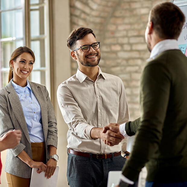 Two business professionals smiling and shaking hands during a meeting, with a woman standing nearby holding a folder.