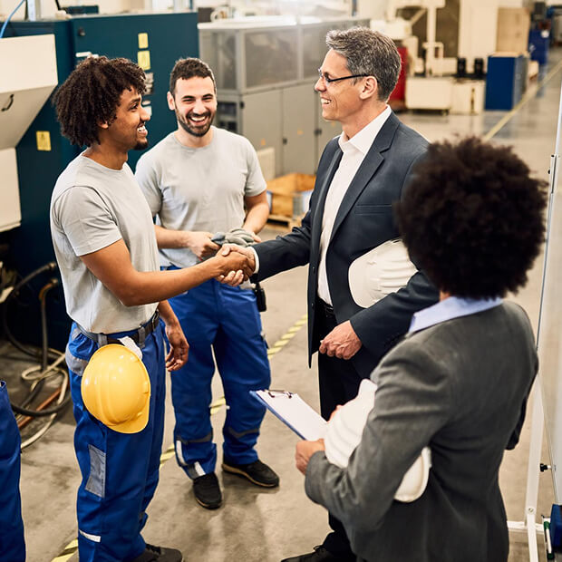 Man in suit shaking hands with a worker in blue pants and gray shirt inside an industrial setting, with two other workers smiling nearby.