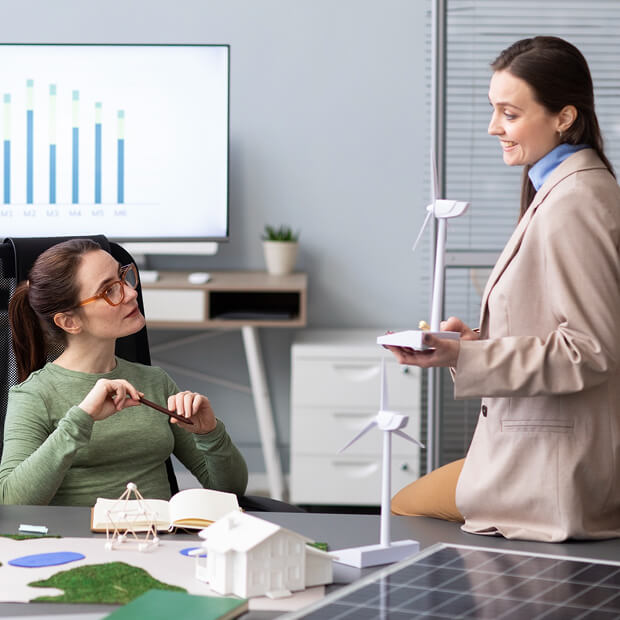 Two women discussing renewable energy models including wind turbines and solar panels in an office setting with a bar graph displayed on a monitor.