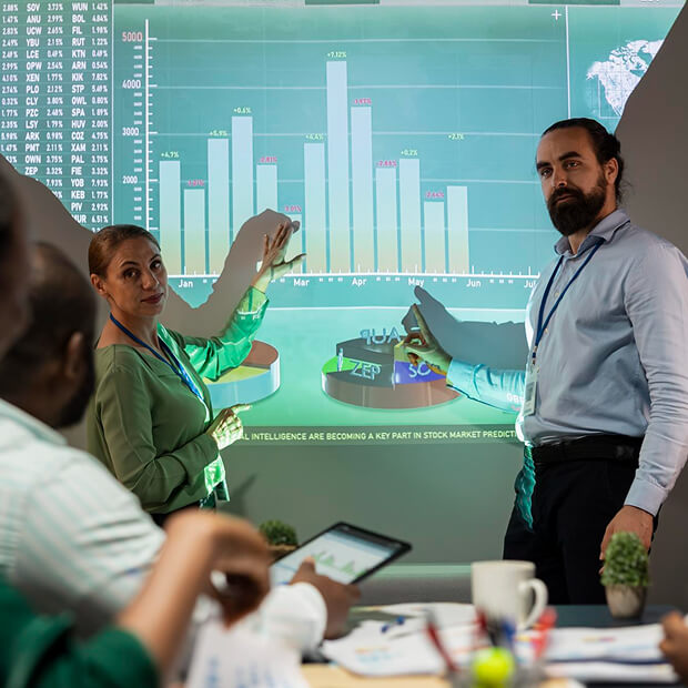 Two professionals presenting and discussing stock market data projected as bar and pie charts in a meeting room.
