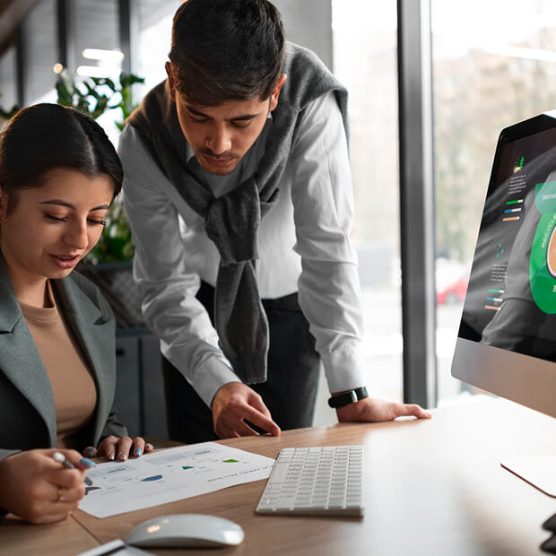 Two colleagues discussing charts and graphs printed on paper at a desk with a computer screen displaying a green circular infographic.