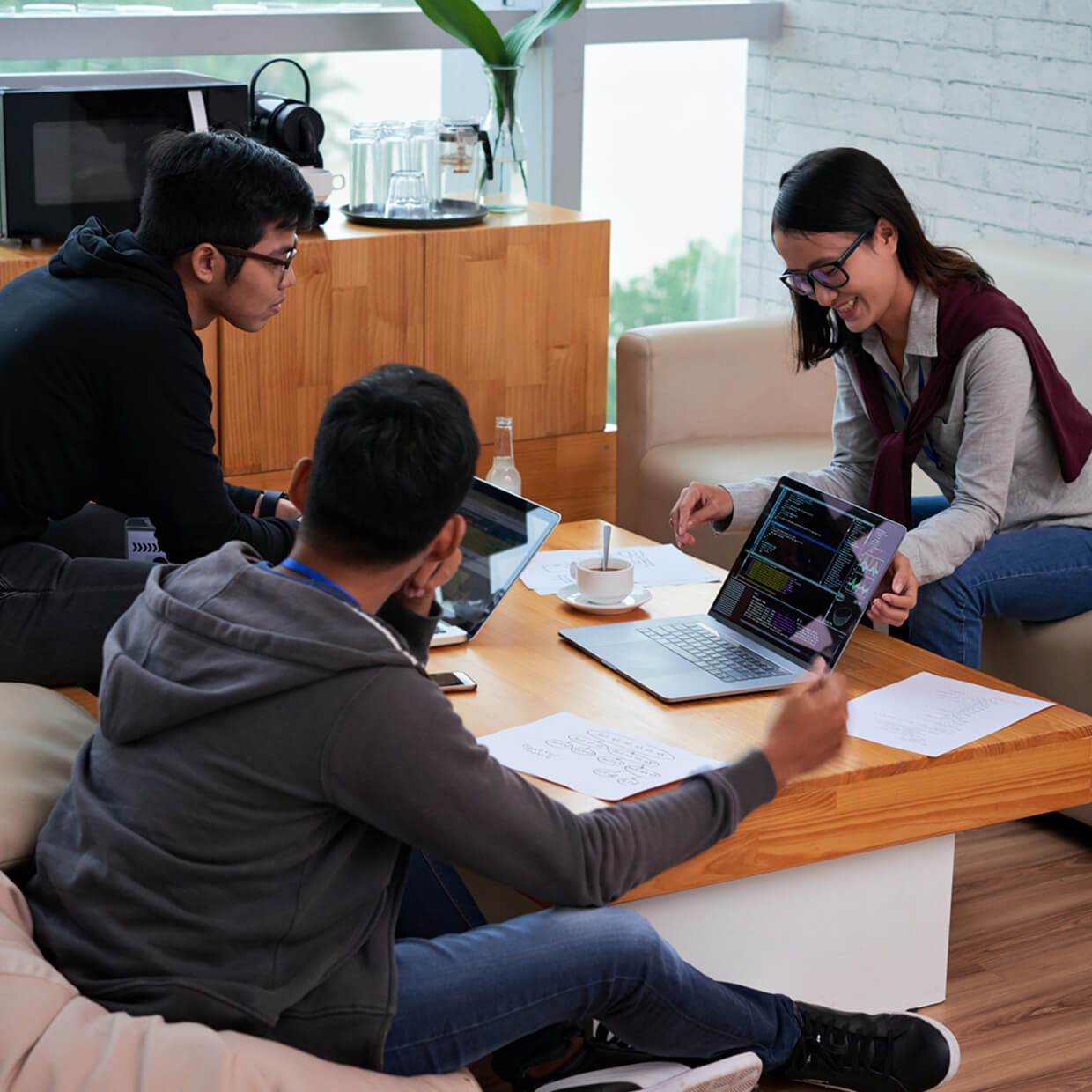 Three young adults collaborate around a wooden table, viewing code on laptops and discussing notes in a casual workspace.