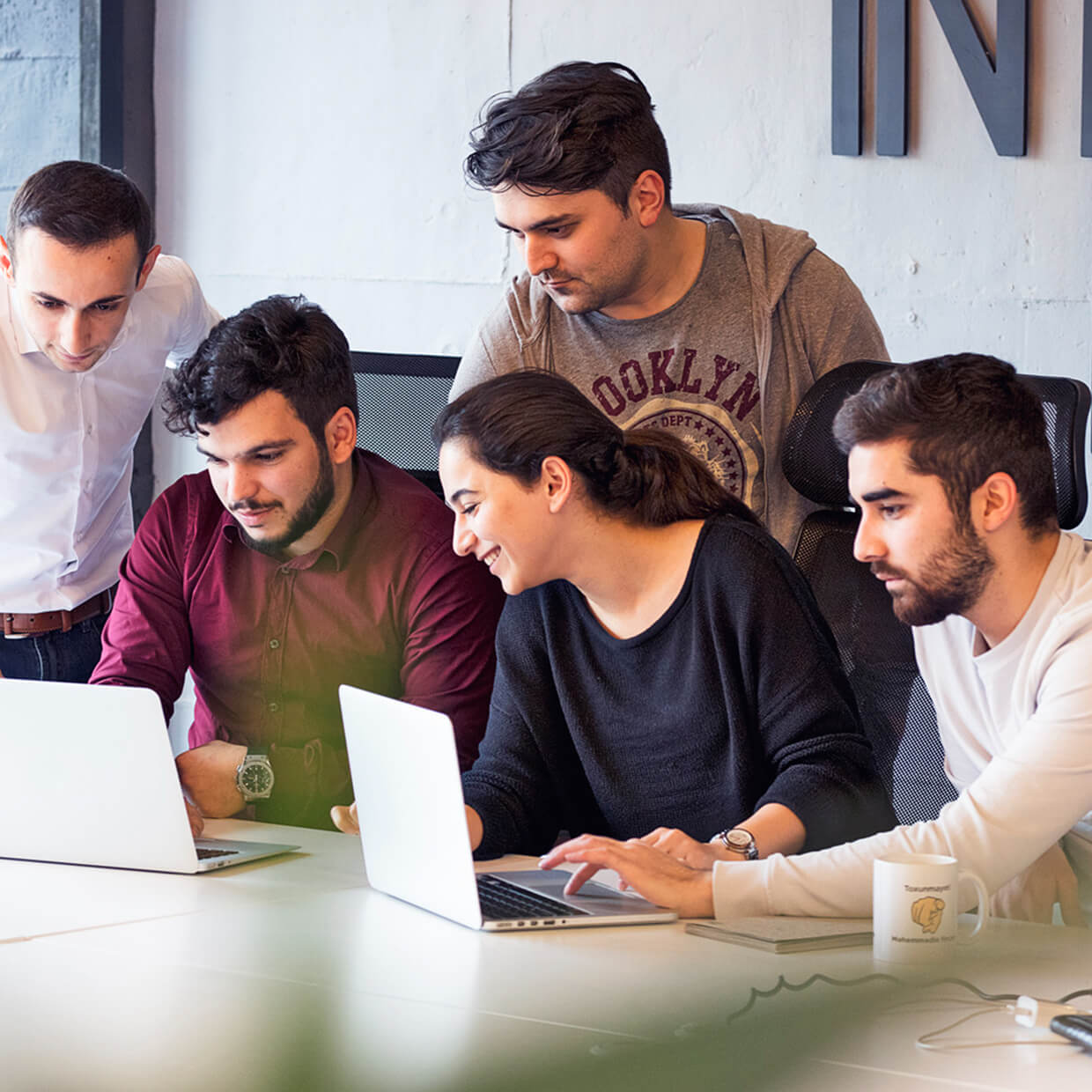 Five young professionals collaborating around a table, working on laptops and discussing work in an office setting.