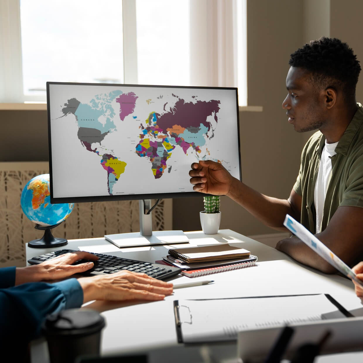 Two people at a desk looking at a computer monitor displaying a colorful world map with a globe and notebooks on the desk.