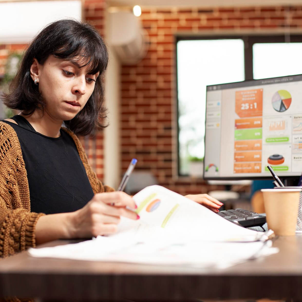 Woman with dark hair reviewing documents while working at a desk with computer displaying charts in an office.