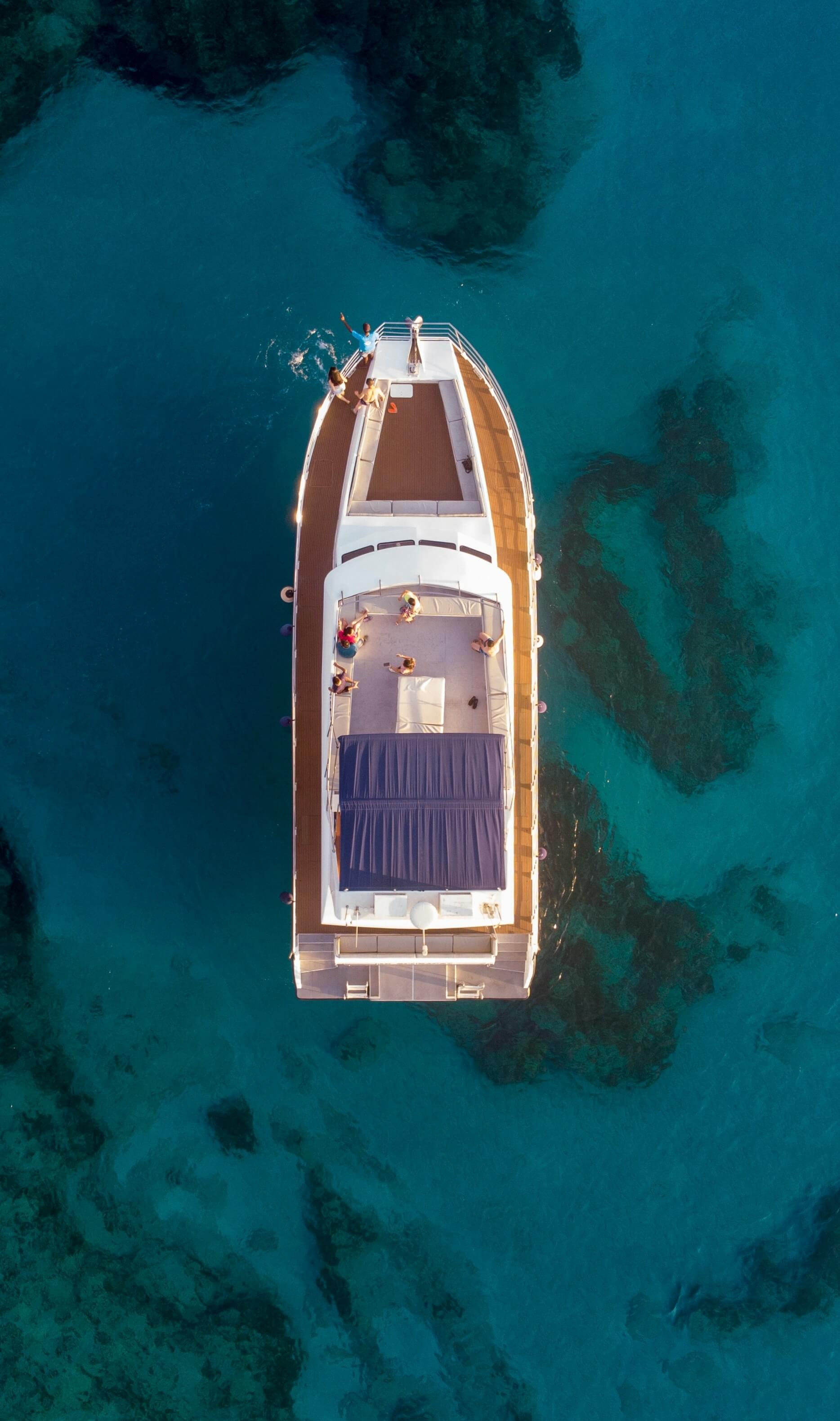 Aerial view of a yacht with several people on deck floating on clear blue water with visible underwater rocks.