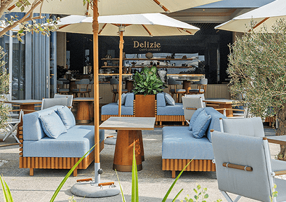 Outdoor seating area with light blue cushioned sofas, wooden tables, and large white umbrellas in front of a café named Delizie.
