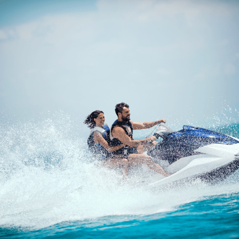 A man and woman wearing life jackets riding a jet ski on blue ocean water with splashing waves.