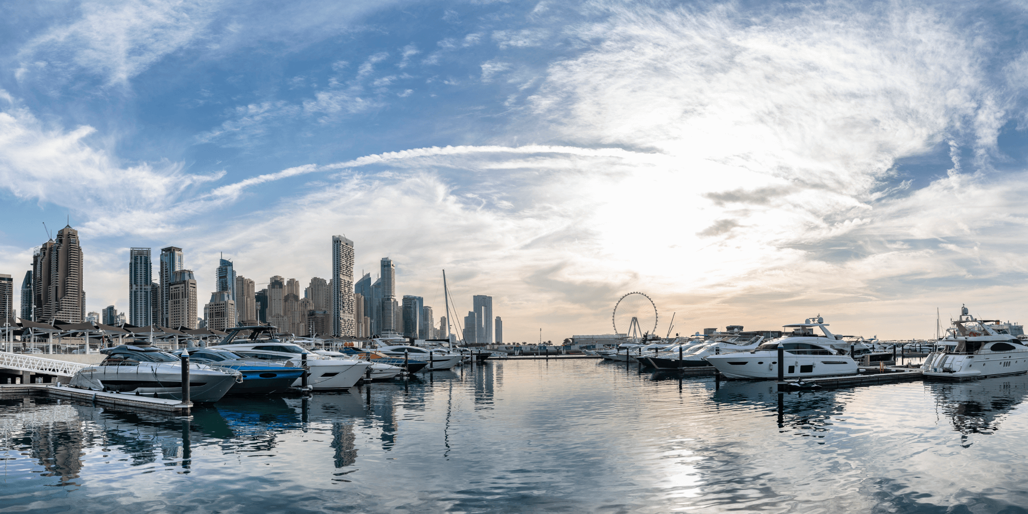 Marina with yachts docked and modern skyscrapers, with a Ferris wheel under a partly cloudy sky.