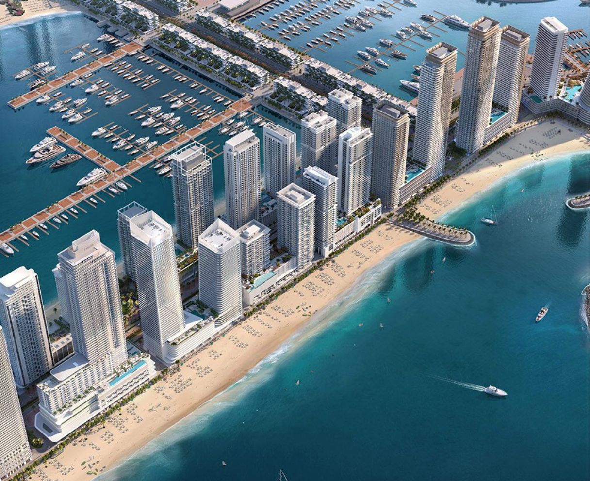 Aerial view of a coastal city with high-rise buildings, sandy beach, marina with yachts, and boats on blue water.