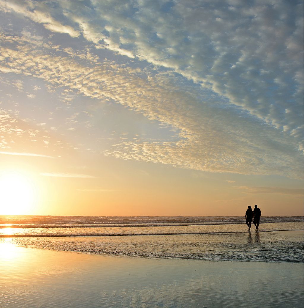 Two people walking on a beach at sunset with a partly cloudy sky overhead.