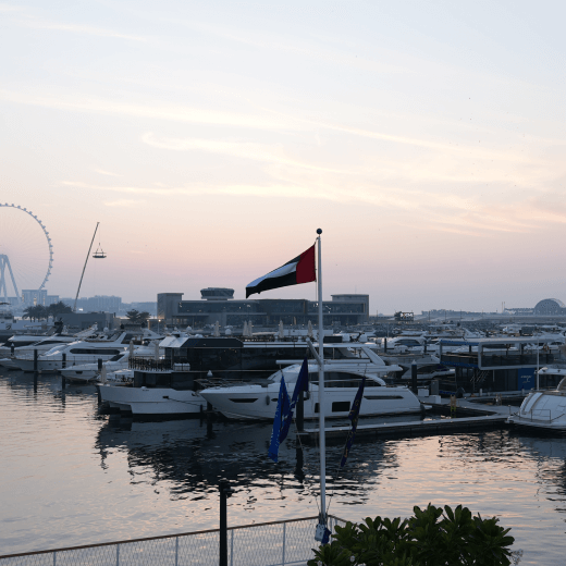 Marina with multiple yachts docked at sunset, with a UAE flag prominently flying and Dubai skyline including a Ferris wheel in the background.