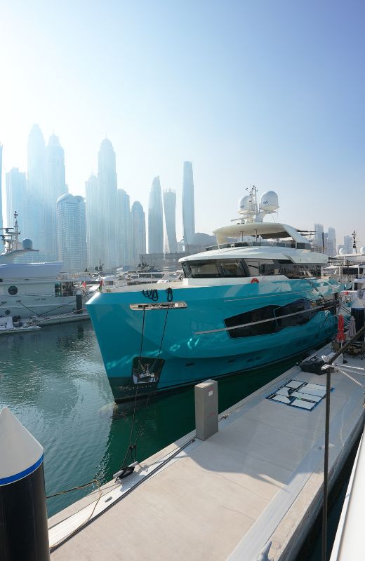 Turquoise yacht docked at a marina with a city skyline in the background under clear sky.