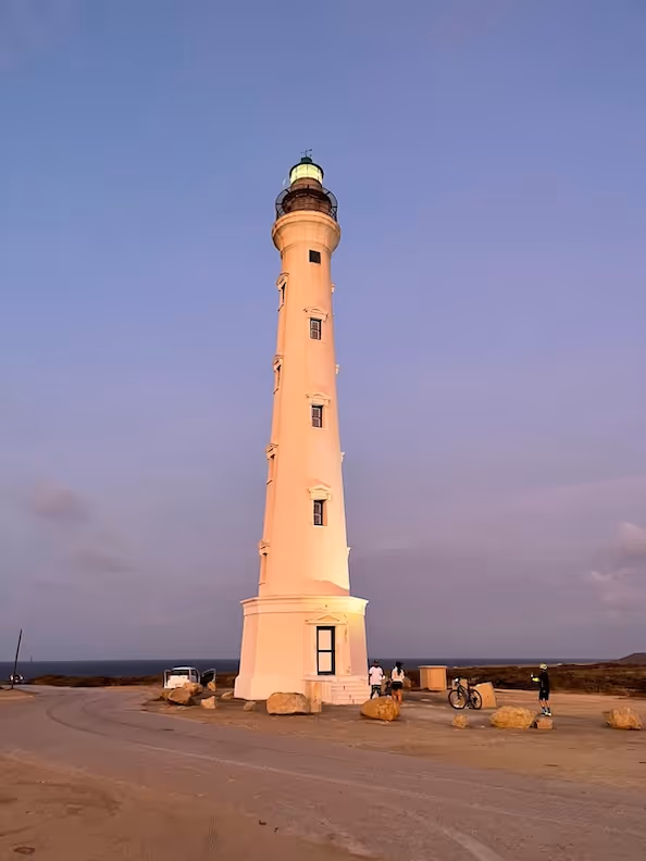 Tall white lighthouse illuminated at dusk with people and bicycles near its base on a sandy area.