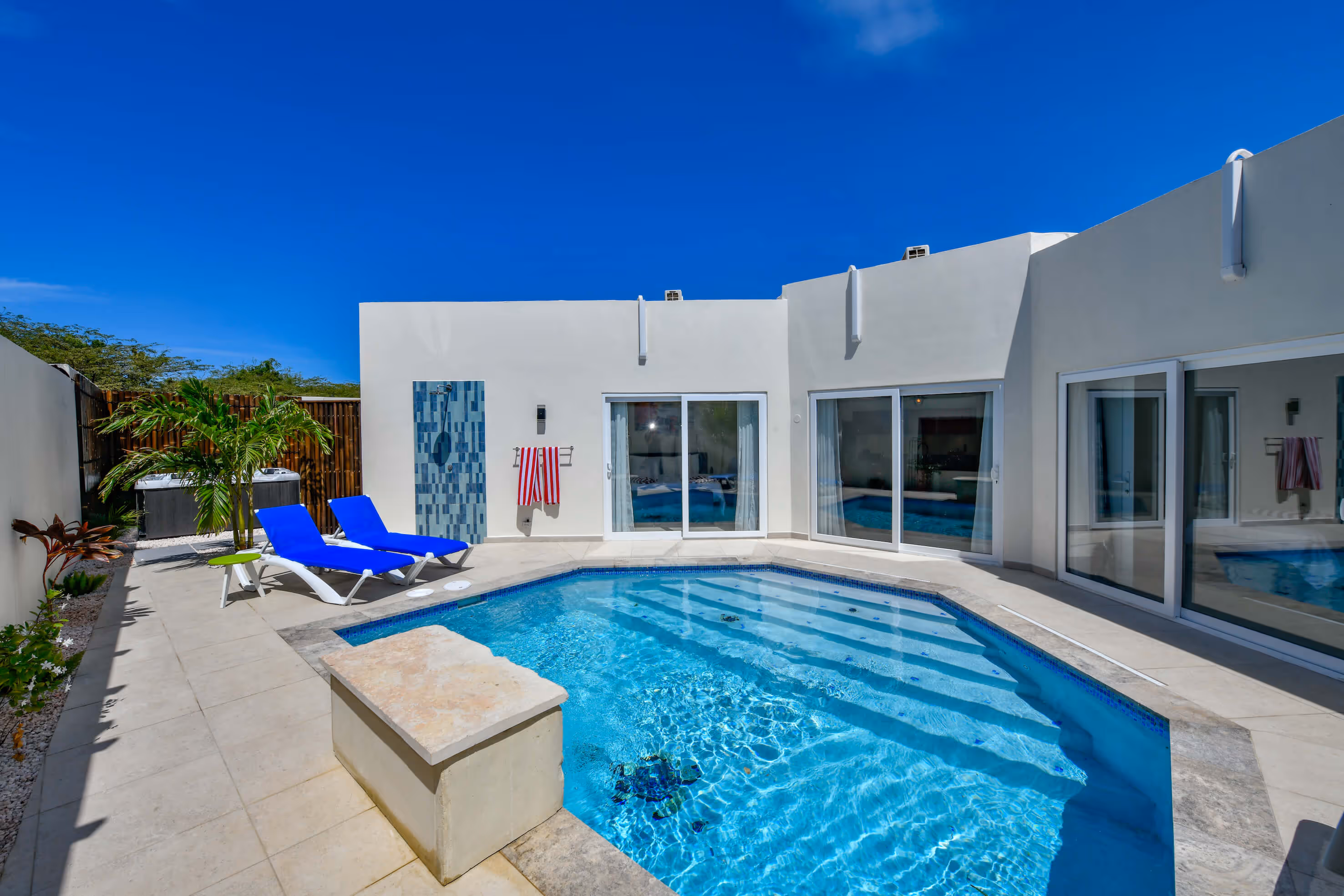 Modern backyard with a clear blue swimming pool, two blue lounge chairs, a small palm tree, and white building walls with sliding glass doors.