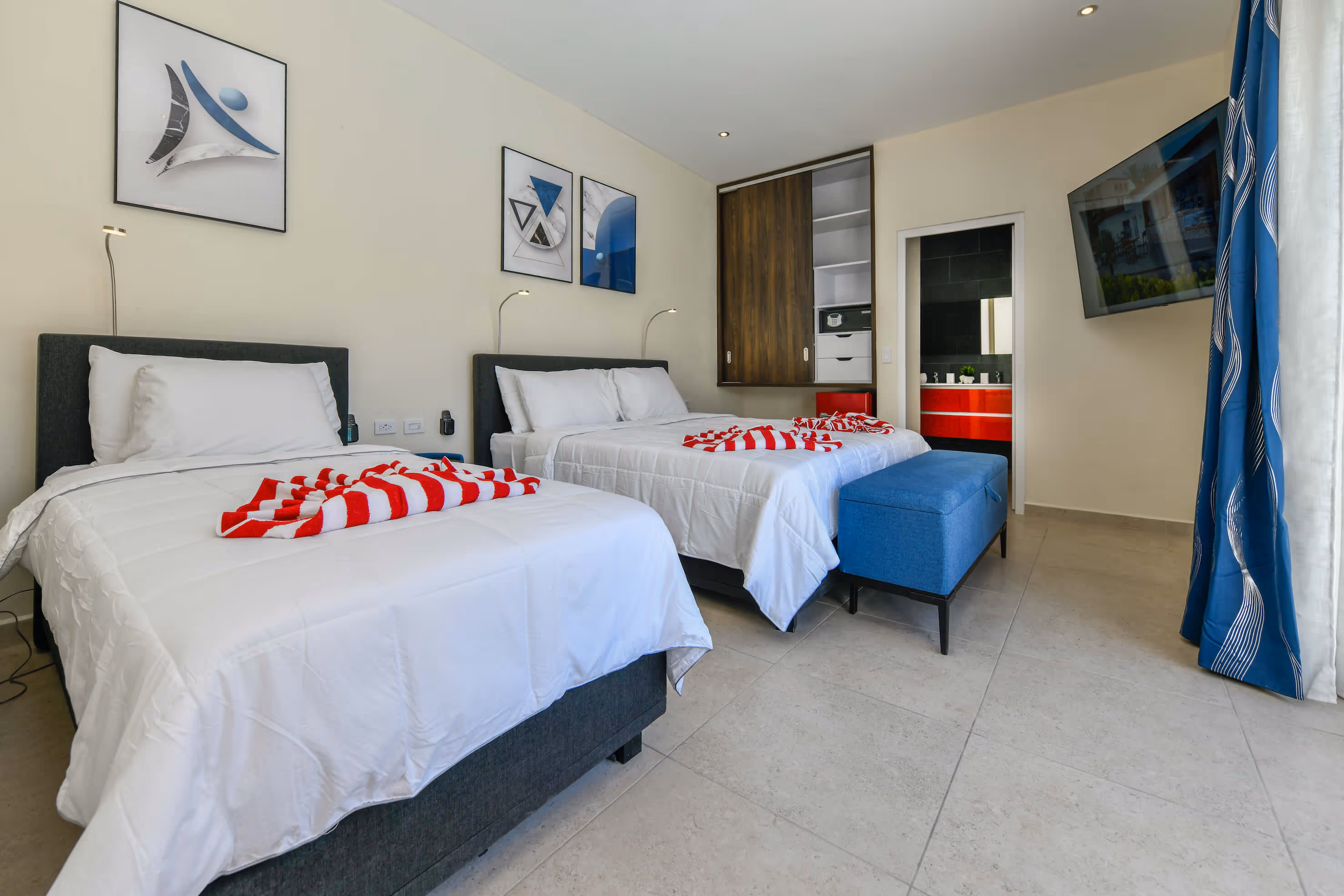 Modern hotel room with two beds covered in white linens and red-striped towels, blue bench, wall-mounted TV, and open closet near a bathroom door.