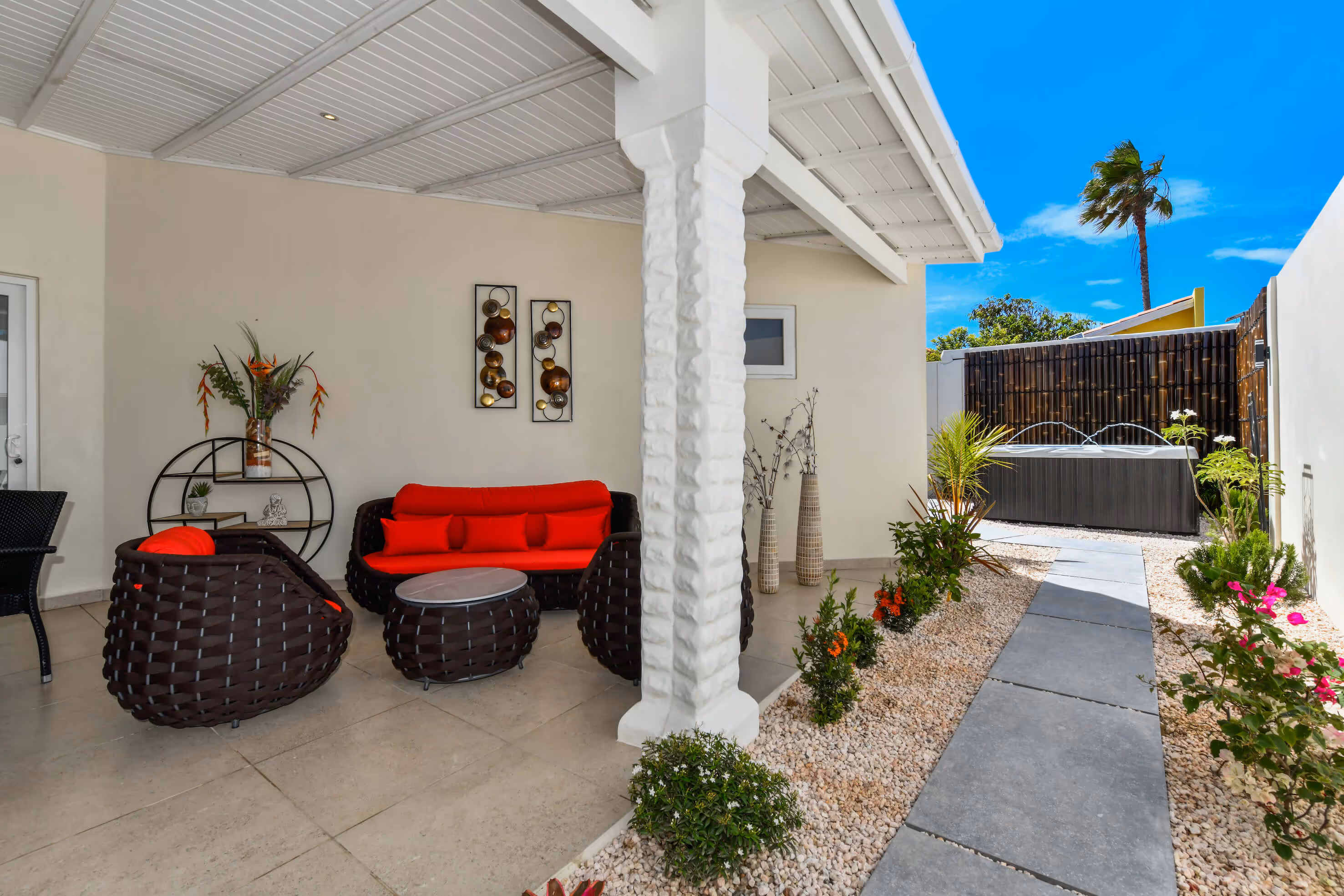 Covered patio with dark wicker furniture featuring red cushions, decorative wall art, and a path leading to a hot tub surrounded by plants under a blue sky.