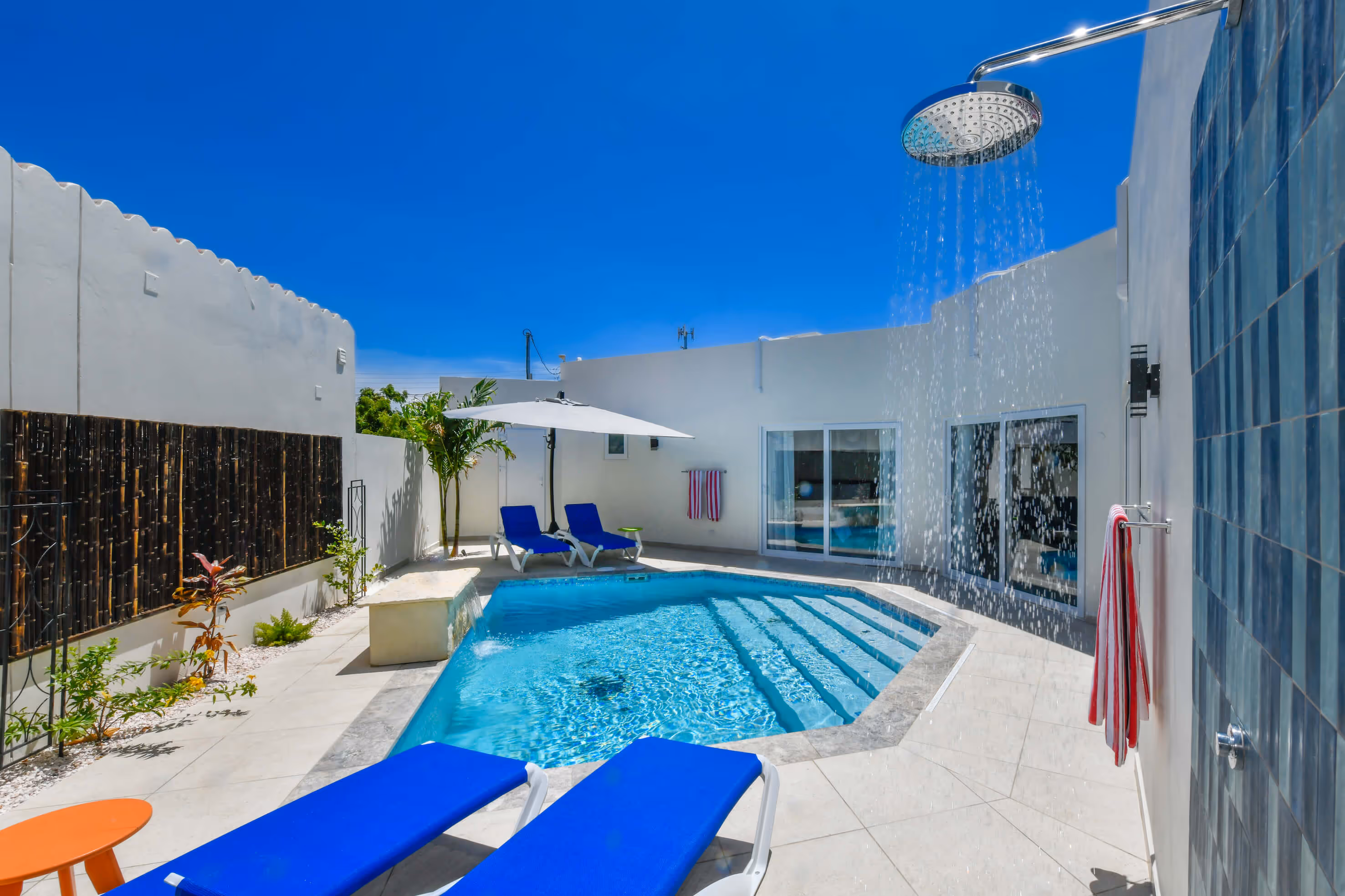Outdoor swimming pool with blue lounge chairs, white umbrella, and a shower spraying water on a sunny day.