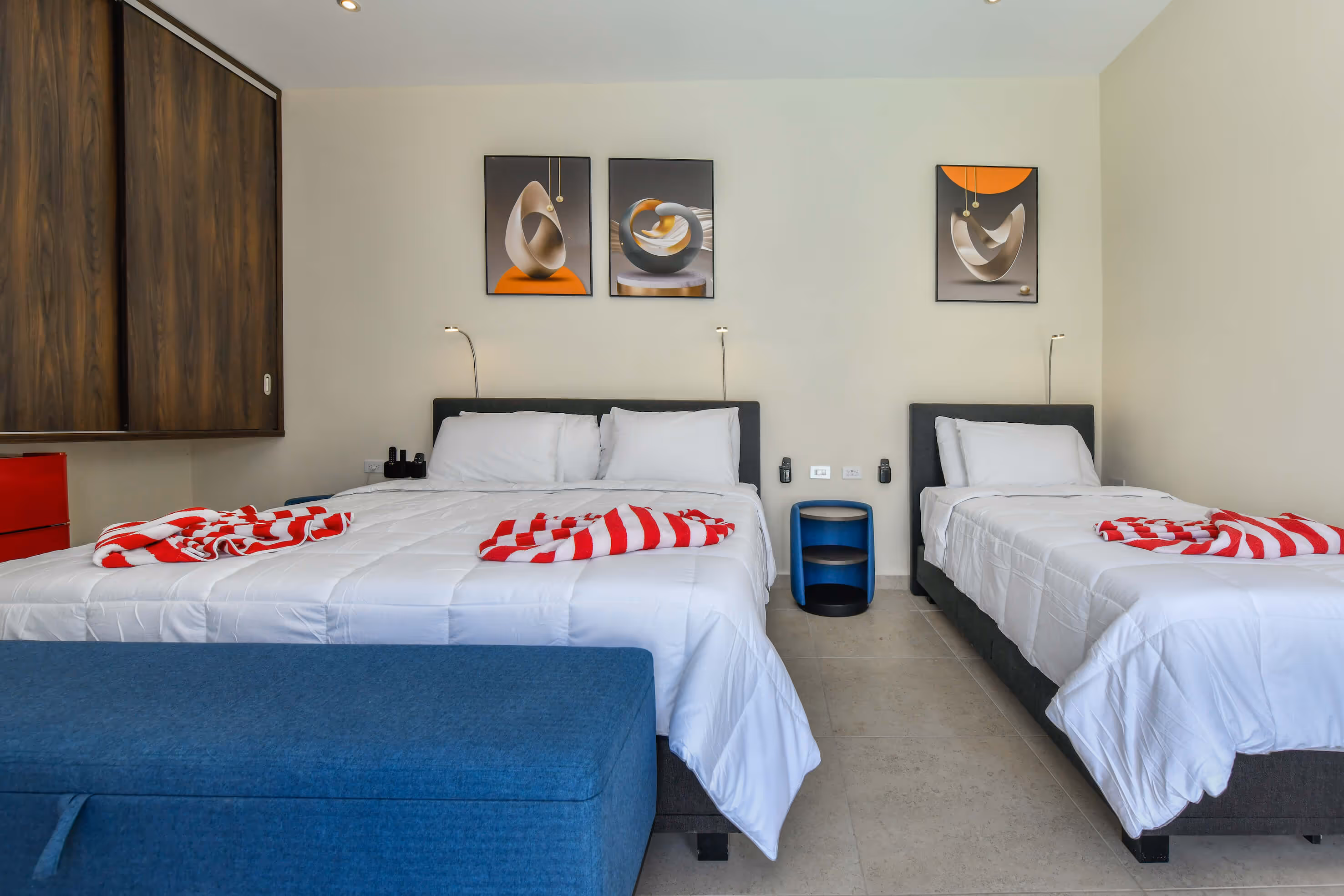 Modern hotel room with one double bed and one single bed with white linens and red-striped towels on them, blue upholstered bench at the foot of the double bed, and three abstract art pieces on the beige wall.