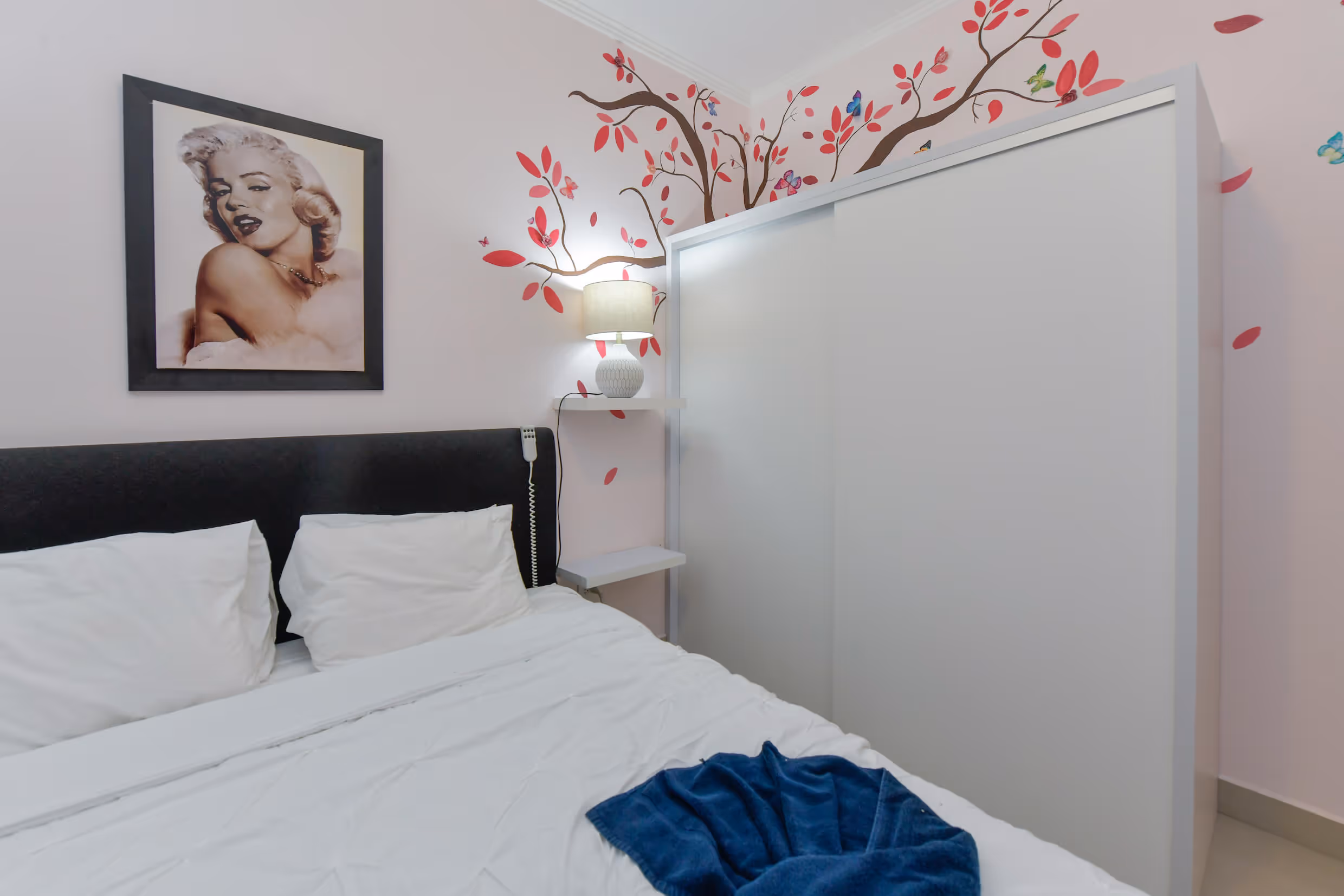 Bedroom with white bed, black headboard, Marilyn Monroe framed photo, lamp on wall shelf, and wardrobe with floral wall decals.