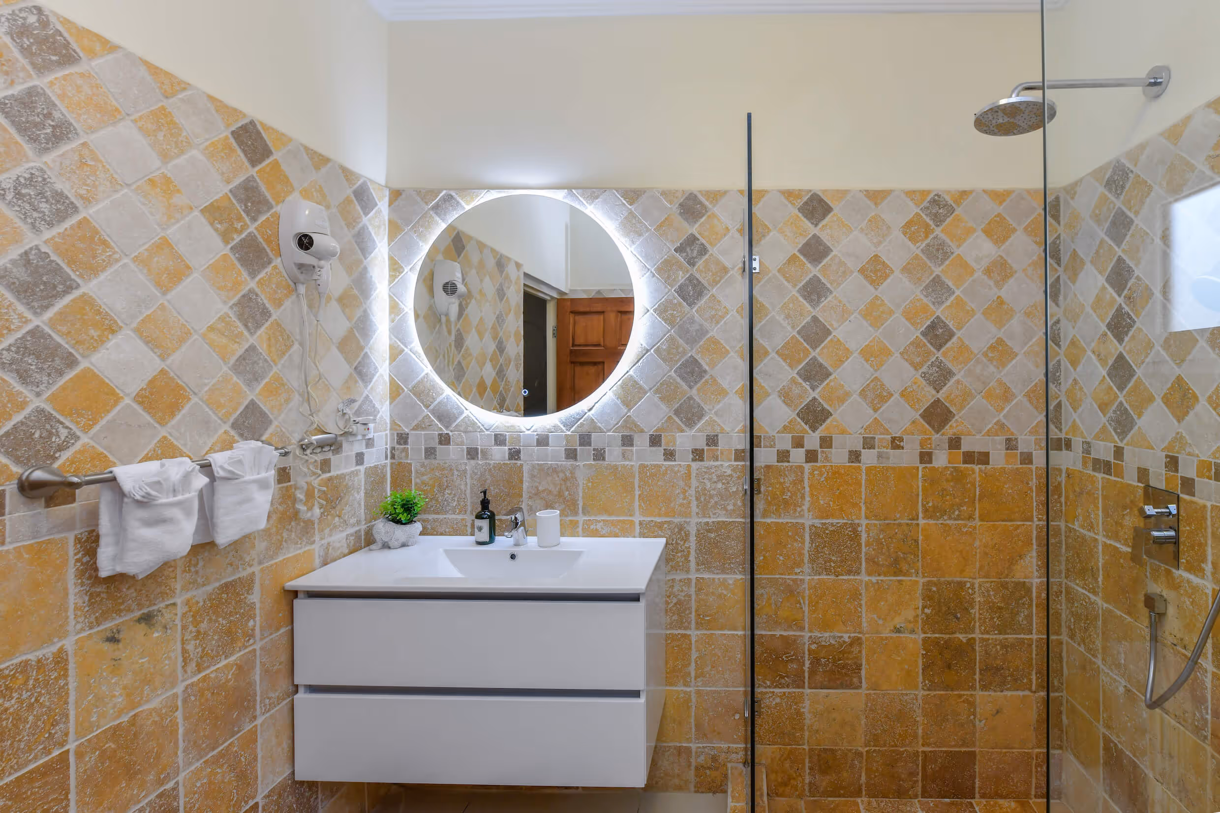 Bathroom with yellow and beige tiled walls, a circular illuminated mirror above a white sink cabinet, a mounted hairdryer, and a glass-enclosed shower area.