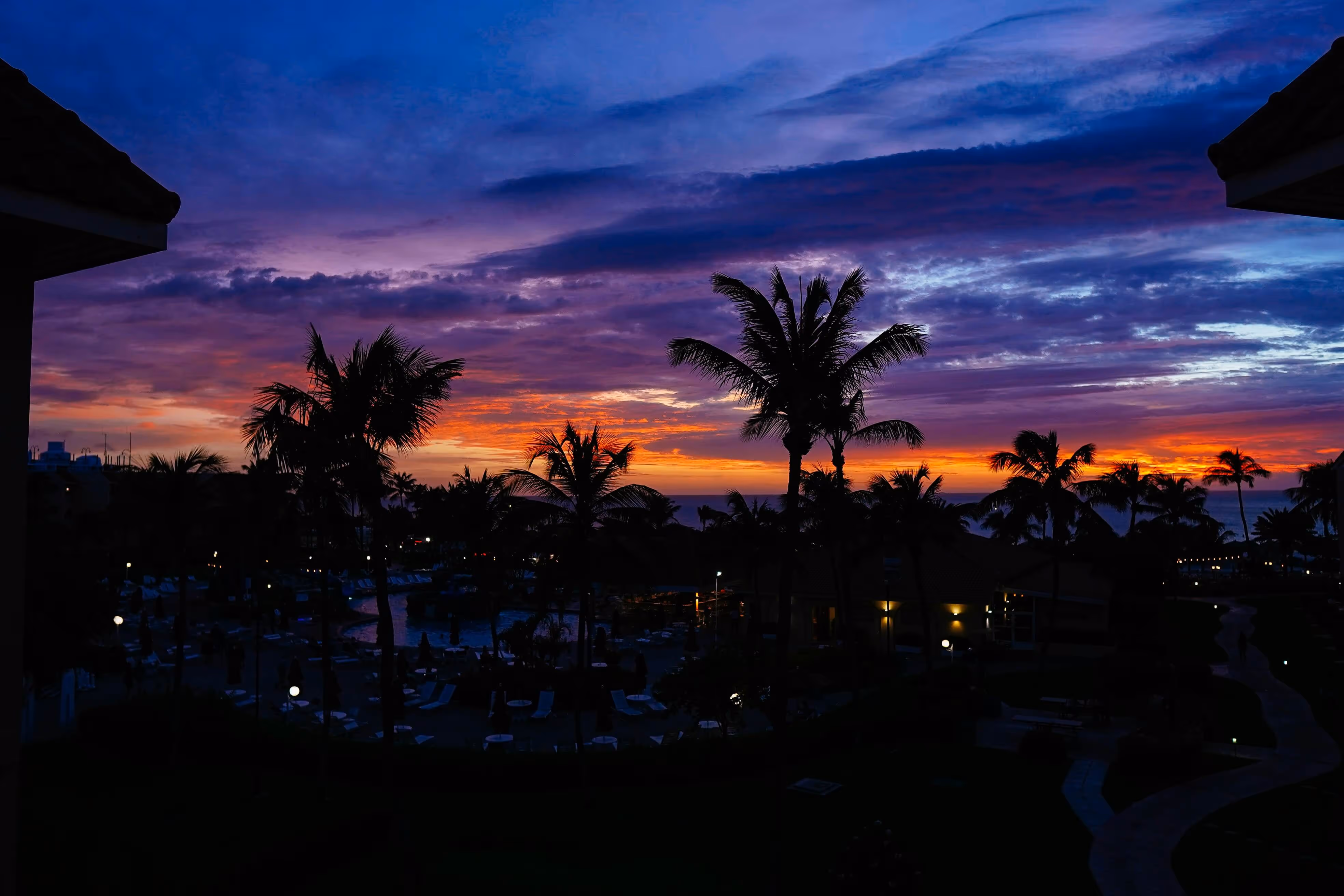Silhouettes of palm trees against a colorful sunset sky over a resort with pool and lounge chairs.
