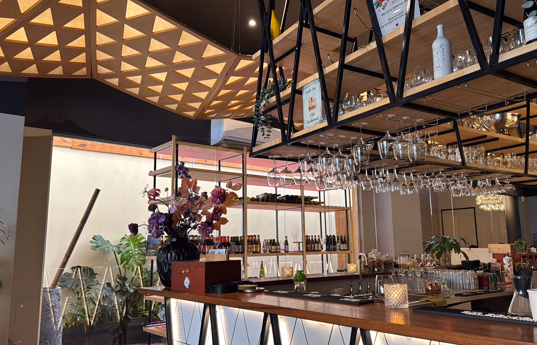 Modern bar interior with wooden counter, hanging wine glasses, shelves with bottles, and a flower arrangement in a black vase.
