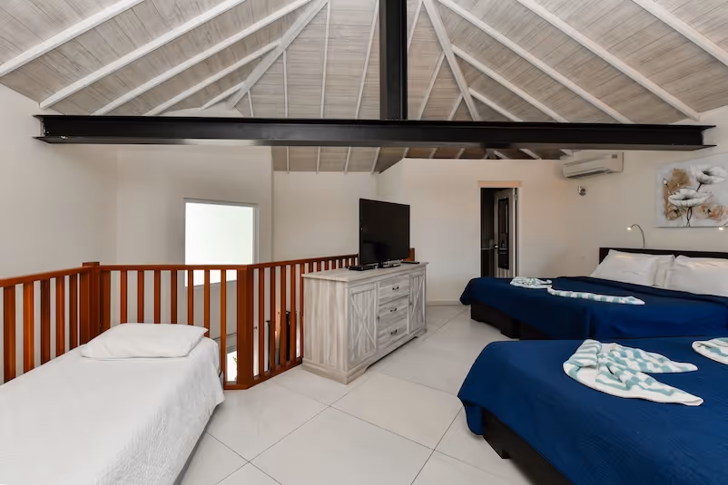 Loft bedroom with white wooden vaulted ceiling, a single bed with white bedding, two double beds with navy blue covers, a TV on a wooden dresser, and wooden railing around stairs.